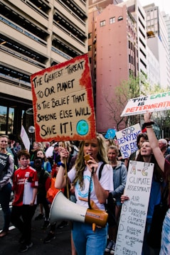 A group of protesters on a city street hold signs with messages about climate change. The central figure carries a megaphone and a sign that reads 'The greatest threat to our planet is the belief that someone else will save it!' Surrounding buildings loom over the crowd, and other demonstrators carry signs with slogans related to environmental action.