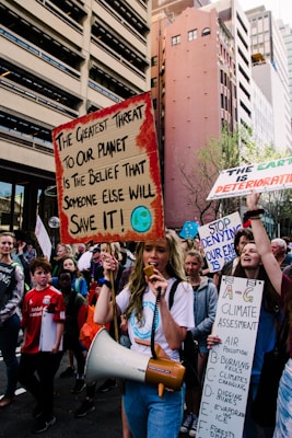 A group of protesters on a city street hold signs with messages about climate change. The central figure carries a megaphone and a sign that reads 'The greatest threat to our planet is the belief that someone else will save it!' Surrounding buildings loom over the crowd, and other demonstrators carry signs with slogans related to environmental action.