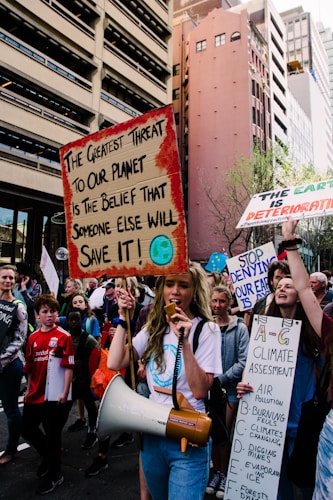 A group of protesters on a city street hold signs with messages about climate change. The central figure carries a megaphone and a sign that reads 'The greatest threat to our planet is the belief that someone else will save it!' Surrounding buildings loom over the crowd, and other demonstrators carry signs with slogans related to environmental action.