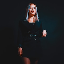 A woman with blonde hair is standing against a dark background. She is wearing a black outfit and is striking a confident pose with one hand placed on her hip.