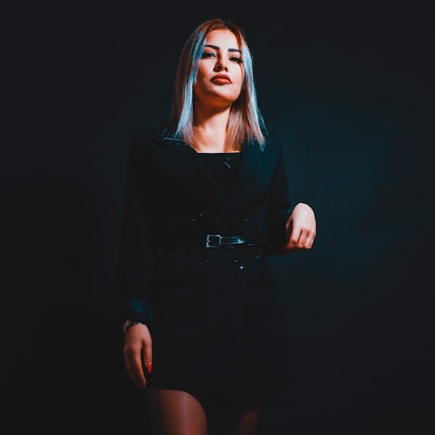A woman with blonde hair is standing against a dark background. She is wearing a black outfit and is striking a confident pose with one hand placed on her hip.