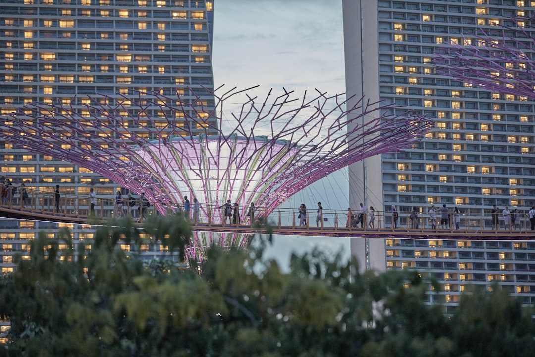 a group of people walking across a bridge in front of tall buildings, People are working by the sky bridge in front of the artificial supertree and hotel Marina Bay Sands