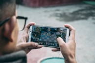 A person is holding a smartphone horizontally, immersed in playing a complex strategy game. The focus is on the game screen, which displays an intricate board setup with various characters and elements.