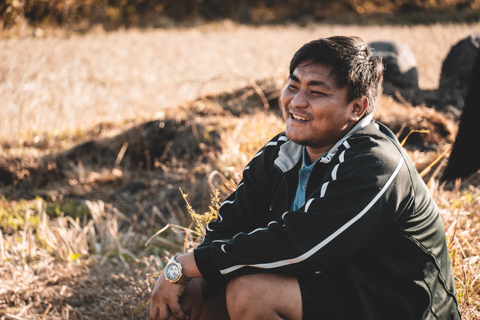 The image shows a young man sitting on the ground, wearing a black jacket with white stripes and shorts. He is smiling at the camera, suggesting he is enjoying his time outdoors. The background features a field with dry grass and trees in the distance, indicating an outdoor setting.