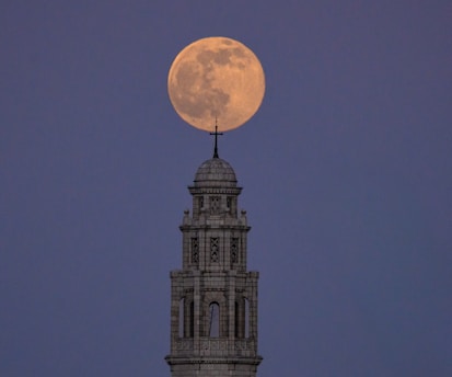 A full moon is positioned directly above the pinnacle of a tall, ornate stone tower. The tower is detailed with intricate architectural features and topped with a small cross. The background is a twilight sky with a serene, dark blue hue.