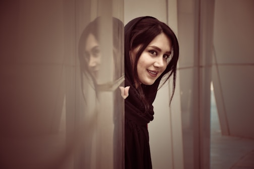 A woman is partially visible, peeking from behind a transparent curtain or panel. Her hair is dark, and she wears a dark outfit, smiling softly as she looks towards the camera. The scene captures a sense of curiosity and gentle intrigue.