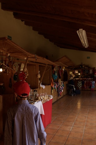 A wooden-paneled indoor market corridor is lined with stalls. A child wearing a red cap and checkered shirt walks along, looking at various handmade crafts, colorful textiles, and packaged goods displayed on tables with red and white covers. The space is warmly lit with overhead fluorescent lights and has a tiled floor.