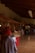 A wooden-paneled indoor market corridor is lined with stalls. A child wearing a red cap and checkered shirt walks along, looking at various handmade crafts, colorful textiles, and packaged goods displayed on tables with red and white covers. The space is warmly lit with overhead fluorescent lights and has a tiled floor.