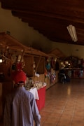 A wooden-paneled indoor market corridor is lined with stalls. A child wearing a red cap and checkered shirt walks along, looking at various handmade crafts, colorful textiles, and packaged goods displayed on tables with red and white covers. The space is warmly lit with overhead fluorescent lights and has a tiled floor.