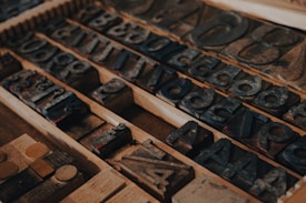 A collection of vintage printing blocks arranged in a drawer, showing various letters and numbers made from dark wood or metal. These blocks are set in rows, showcasing the intricacies of old-school typography used in traditional printing presses.