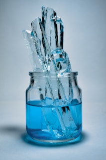 Several tall, clear ice chunks are standing upright in a glass jar filled with vibrant blue liquid. The scene is set against a soft, neutral background, creating a cool and refreshing visual effect.