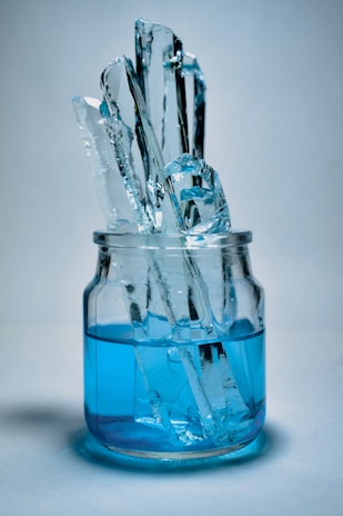 Several tall, clear ice chunks are standing upright in a glass jar filled with vibrant blue liquid. The scene is set against a soft, neutral background, creating a cool and refreshing visual effect.