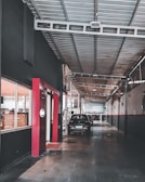 A long, industrial corridor inside a workshop, with a high ceiling supported by metal beams. Cars are parked along the pathway, and windows on the left show interiors of well-lit rooms. The walls are painted dark with red accents, and the floor is made of concrete.