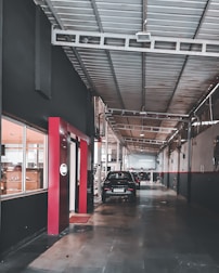 A long, industrial corridor inside a workshop, with a high ceiling supported by metal beams. Cars are parked along the pathway, and windows on the left show interiors of well-lit rooms. The walls are painted dark with red accents, and the floor is made of concrete.