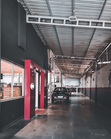 A long, industrial corridor inside a workshop, with a high ceiling supported by metal beams. Cars are parked along the pathway, and windows on the left show interiors of well-lit rooms. The walls are painted dark with red accents, and the floor is made of concrete.
