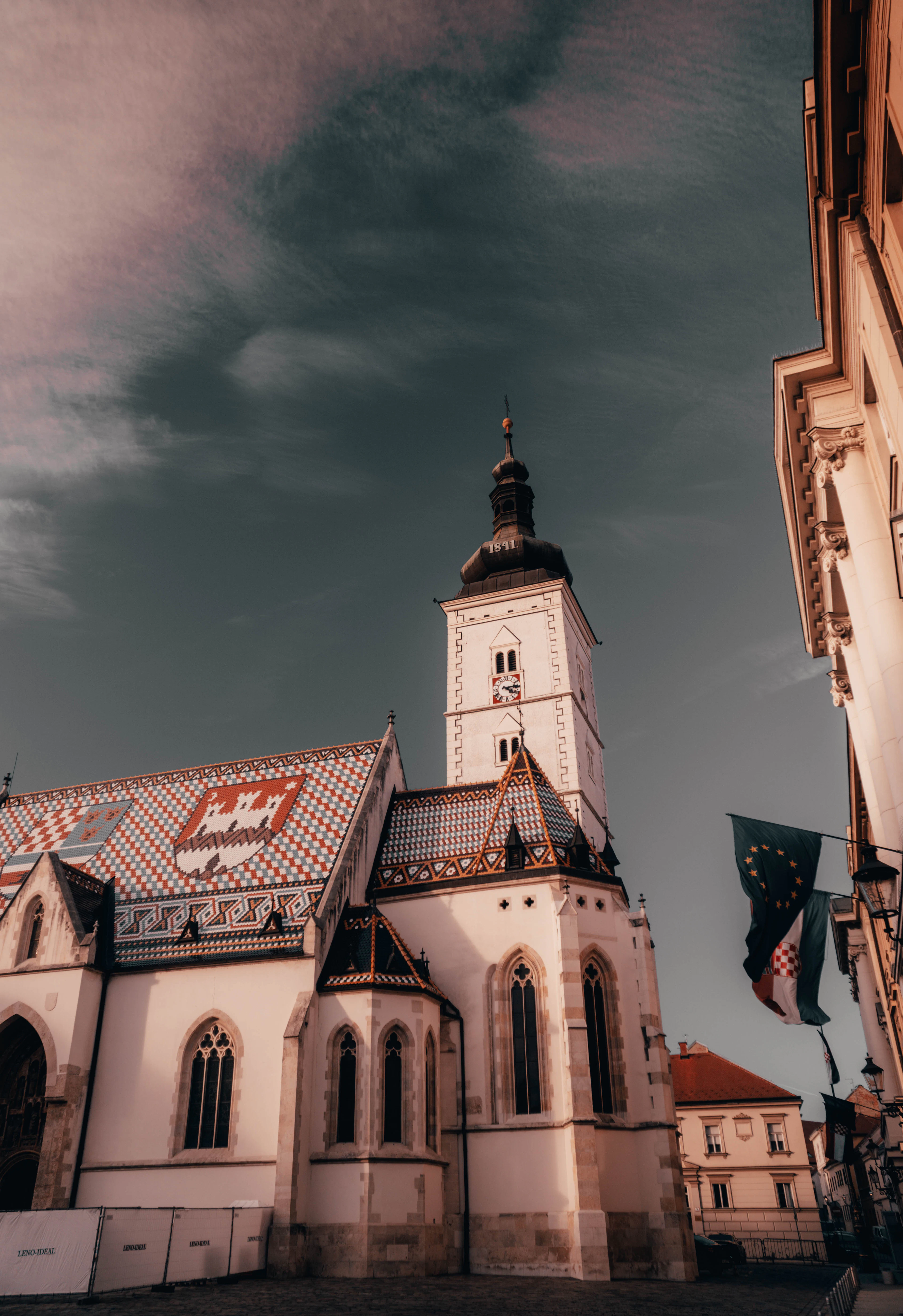 St. Mark's Church with its patterned tiled roof and spire lit warmly against a dark, stormy sky, flags flying beside it.