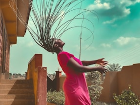 A person with braided hair arches their head back, causing the braids to fan out dramatically. They are wearing a bright pink dress, and the setting appears to be an outdoor area with steps, a wall, plants, and a partly cloudy sky. The lighting is natural, creating a shadowed effect under the eaves of the structure.