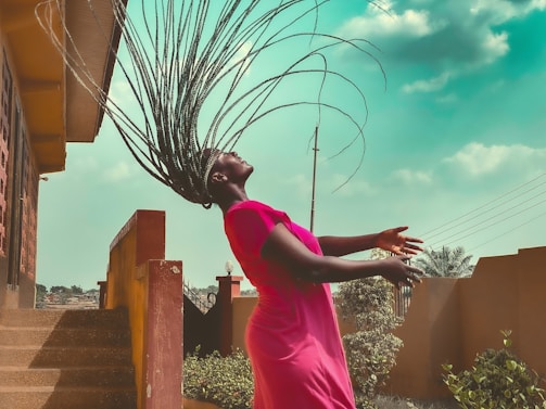 A person with braided hair arches their head back, causing the braids to fan out dramatically. They are wearing a bright pink dress, and the setting appears to be an outdoor area with steps, a wall, plants, and a partly cloudy sky. The lighting is natural, creating a shadowed effect under the eaves of the structure.