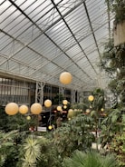 A large indoor atrium filled with lush green plants and trees under a glass-paneled ceiling. Several spherical, warm-colored lanterns are suspended from the ceiling, casting a soft glow over the foliage. The architecture features a modern, industrial design with visible metal framework and large windows.