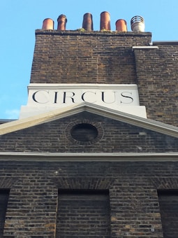 A brick building facade with the word 'CIRCUS' displayed prominently beneath a row of chimneys. The structure features distinct brickwork patterns and a circular vent above the windows. The sky is clear and blue in the background.