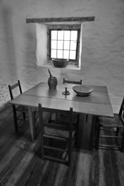 A rustic interior featuring a wooden table with four chairs in a dimly lit room. On the table are various objects including a woven basket, a candlestick holder, and a jug. A window with multiple panes lets in natural light, casting shadows across the wooden floor.