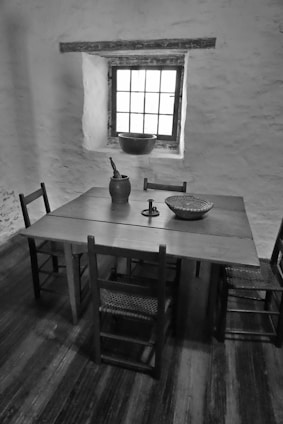 A rustic interior featuring a wooden table with four chairs in a dimly lit room. On the table are various objects including a woven basket, a candlestick holder, and a jug. A window with multiple panes lets in natural light, casting shadows across the wooden floor.