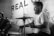 A bearded man energetically plays the drums in an intimate indoor setting, wearing a casual t-shirt. The background features a wall with a sign that says 'REAL' and some decorative bunting.