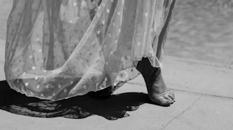A black and white photograph captures bare feet walking gracefully on a concrete surface. The lower part of a sheer, polka-dotted dress sways with movement, casting soft shadows.