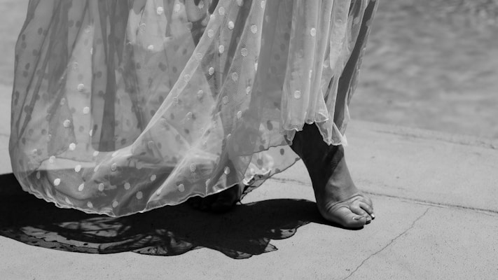 A black and white photograph captures bare feet walking gracefully on a concrete surface. The lower part of a sheer, polka-dotted dress sways with movement, casting soft shadows.