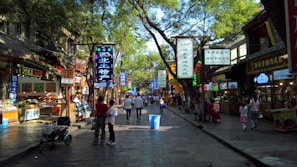 A bustling street market scene filled with various shop signs in Chinese characters. People are walking along the stone-paved path lined with trees, creating a blend of shadow and light. Stalls on both sides display food and goods, with some vendors attending to customers.