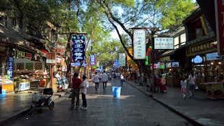 A bustling street market scene filled with various shop signs in Chinese characters. People are walking along the stone-paved path lined with trees, creating a blend of shadow and light. Stalls on both sides display food and goods, with some vendors attending to customers.