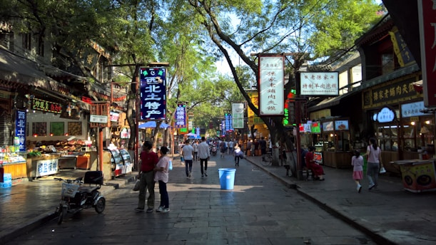 A bustling street market scene filled with various shop signs in Chinese characters. People are walking along the stone-paved path lined with trees, creating a blend of shadow and light. Stalls on both sides display food and goods, with some vendors attending to customers.