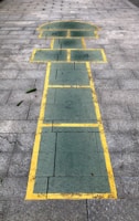 A hopscotch game drawn on a paved surface with green rectangles and squares bordered by yellow lines. The surface consists of grey brick tiles. The layout includes numbered sections from 1 to 4, and parts of the drawing resemble the shape of a house or a figure.