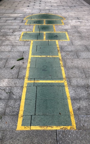 A hopscotch game drawn on a paved surface with green rectangles and squares bordered by yellow lines. The surface consists of grey brick tiles. The layout includes numbered sections from 1 to 4, and parts of the drawing resemble the shape of a house or a figure.