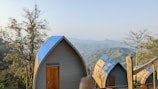 Several small A-frame cabins with blue roofs are situated on a hillside, surrounded by trees and overlooking a scenic view of distant, rolling hills under a clear sky.