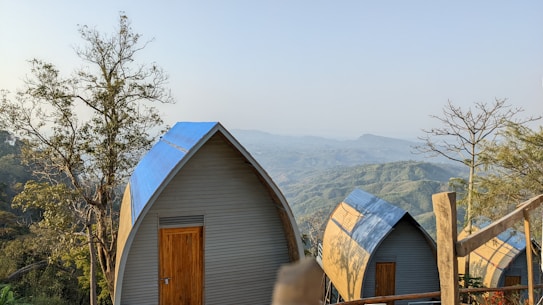 Several small A-frame cabins with blue roofs are situated on a hillside, surrounded by trees and overlooking a scenic view of distant, rolling hills under a clear sky.