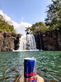A can of Red Bull energy drink is held in the foreground against a backdrop of a waterfall cascading into a clear, green pool. The waterfall is surrounded by rocky cliffs and lush greenery. The sky is clear with a few clouds visible.