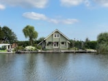 A charming green house with a steep roof is situated on a waterfront. The house is surrounded by lush greenery with trees and bushes, and a wooden deck extends over the water. The scene includes a clear sky with light clouds and a peaceful body of water in the foreground.