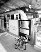 A bicycle is folded up and placed next to the entrance of a modern train at a railway station. The signage on the doors features text in an Indonesian language along with a website link. Overhead beams and reflective surfaces create an industrial atmosphere.