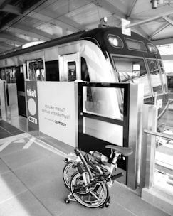 A bicycle is folded up and placed next to the entrance of a modern train at a railway station. The signage on the doors features text in an Indonesian language along with a website link. Overhead beams and reflective surfaces create an industrial atmosphere.