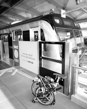 A bicycle is folded up and placed next to the entrance of a modern train at a railway station. The signage on the doors features text in an Indonesian language along with a website link. Overhead beams and reflective surfaces create an industrial atmosphere.