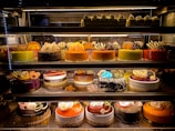 An assortment of colorful and intricately decorated cakes are displayed on shelves in a bakery. The cakes vary in flavor and design, with layers of frosting, fruit, chocolate, and decorative toppings prominently featured. Some of the cakes are round while others have more unique shapes, all presented in an enticing and artistic manner.
