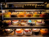 An assortment of colorful and intricately decorated cakes are displayed on shelves in a bakery. The cakes vary in flavor and design, with layers of frosting, fruit, chocolate, and decorative toppings prominently featured. Some of the cakes are round while others have more unique shapes, all presented in an enticing and artistic manner.