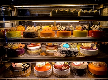An assortment of colorful and intricately decorated cakes are displayed on shelves in a bakery. The cakes vary in flavor and design, with layers of frosting, fruit, chocolate, and decorative toppings prominently featured. Some of the cakes are round while others have more unique shapes, all presented in an enticing and artistic manner.
