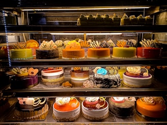 An assortment of colorful and intricately decorated cakes are displayed on shelves in a bakery. The cakes vary in flavor and design, with layers of frosting, fruit, chocolate, and decorative toppings prominently featured. Some of the cakes are round while others have more unique shapes, all presented in an enticing and artistic manner.
