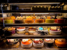 An assortment of colorful and intricately decorated cakes are displayed on shelves in a bakery. The cakes vary in flavor and design, with layers of frosting, fruit, chocolate, and decorative toppings prominently featured. Some of the cakes are round while others have more unique shapes, all presented in an enticing and artistic manner.