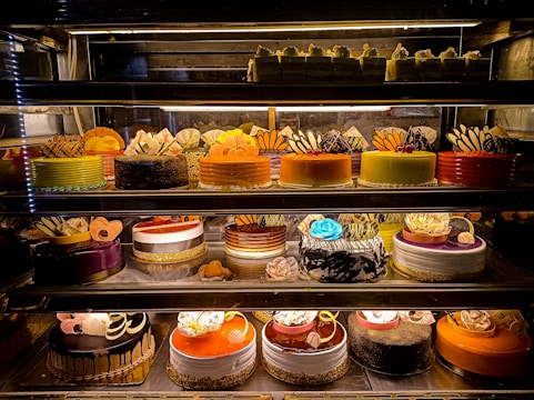 An assortment of colorful and intricately decorated cakes are displayed on shelves in a bakery. The cakes vary in flavor and design, with layers of frosting, fruit, chocolate, and decorative toppings prominently featured. Some of the cakes are round while others have more unique shapes, all presented in an enticing and artistic manner.