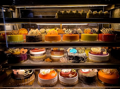 An assortment of colorful and intricately decorated cakes are displayed on shelves in a bakery. The cakes vary in flavor and design, with layers of frosting, fruit, chocolate, and decorative toppings prominently featured. Some of the cakes are round while others have more unique shapes, all presented in an enticing and artistic manner.