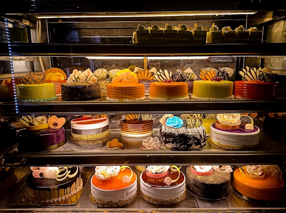 An assortment of colorful and intricately decorated cakes are displayed on shelves in a bakery. The cakes vary in flavor and design, with layers of frosting, fruit, chocolate, and decorative toppings prominently featured. Some of the cakes are round while others have more unique shapes, all presented in an enticing and artistic manner.