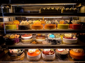 An assortment of colorful and intricately decorated cakes are displayed on shelves in a bakery. The cakes vary in flavor and design, with layers of frosting, fruit, chocolate, and decorative toppings prominently featured. Some of the cakes are round while others have more unique shapes, all presented in an enticing and artistic manner.
